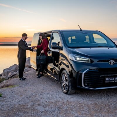 Driver greeting passengers next to a Toyota Proace Verso transfer van parked by the coast.